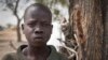 In this photo taken Saturday, Feb. 15, 2014, Ajing Abiik, 12, who fled from his hometown of Bor when the fighting broke out in December, stands next to the tree where he and his two brothers sleep in Minkaman IDP camp, Awerial County, in South Sudan. The fighting in the world’s newest country has left thousands of its youngest citizens either orphans or separated from their parents, increasing their vulnerability to sickness, malnutrition and recruitment by warring groups as child soldiers. (AP Photo/Mackenzie Knowles-Coursin)