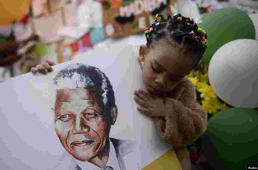 Precious Mali, seorang anak berusia dua tahun memegang gambar Nelson Mandela bersama sejumlah orang lainnya yang berdoa untuk kesembuhan mantan Presiden Afrika Selatan itu, di luar rumah sakit jantung Medi-Clinic tempat Mandela dirawat di Pretoria.