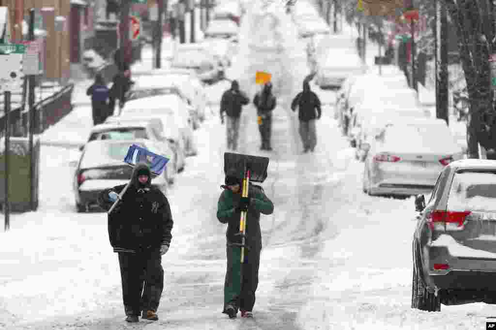 Para pria berjalan membawa pengki di tengah jalanan yang ditutupi salju setelah badai salju di Hoboken, New Jersey (27/1). (AP/Julio Cortez)