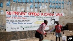 Vijana bawili wanauza njugu mbele ya tangazo linalowashauri watu kuchukua tahadhari dhidi kupata maambukizi mapya ya virusi vya corona, katika eneo la Kibera ambalo ni makazi ya watu yasiorasimishwa, Nairobi, Kenya, Alhamisi, Aprili 2, 2020.